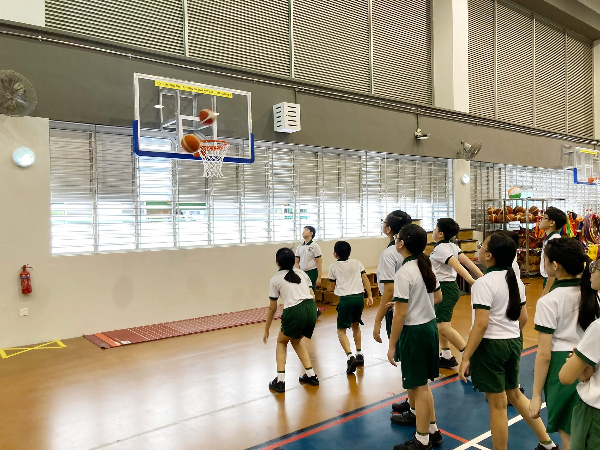 Students gather beneath a basketball hoop, watching as a basketball drops through the net during a shooting activity in the indoor sports hall.